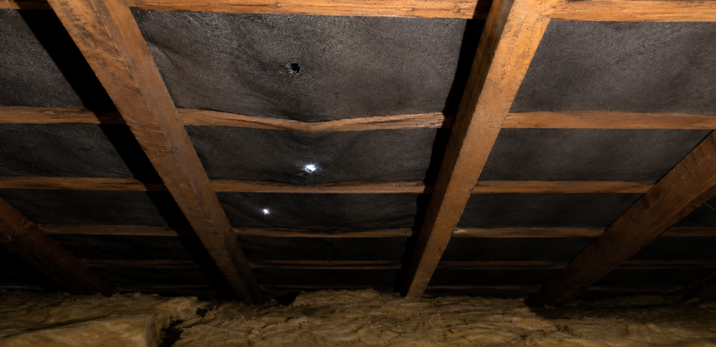 “Interior view of a dark attic roof structure with exposed wooden rafters, black roofing underlay, visible holes letting in daylight, and insulation material along the floor.”