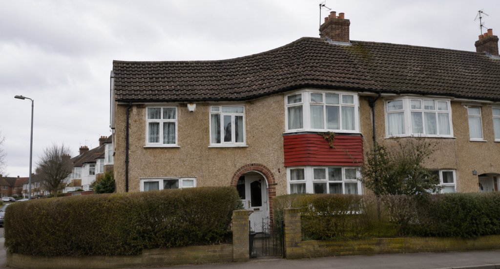 “Traditional semi-detached suburban house with a tiled roof, bay windows, and front hedge on a residential street under an overcast sky.