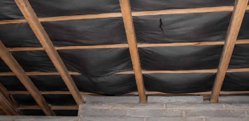 “Interior attic roof structure with exposed timber rafters and torn black roofing membrane showing signs of sagging and potential roof damage.”