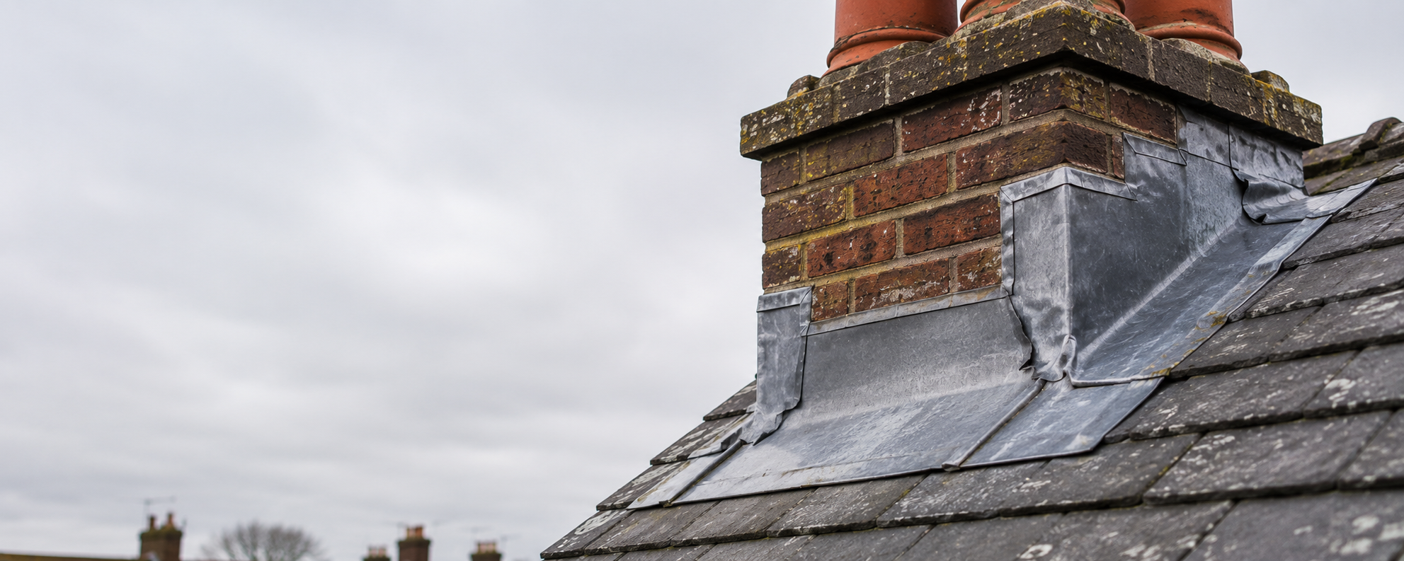 Weathered lead flashing around a brick chimney showing signs of deterioration on a slate roof