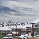 Snow-covered suburban houses with white rooftops line a residential neighborhood under a cloudy sky, hinting at the importance of roof lifespan in enduring harsh winters. Green shrubs and fences are visible in the foreground.
