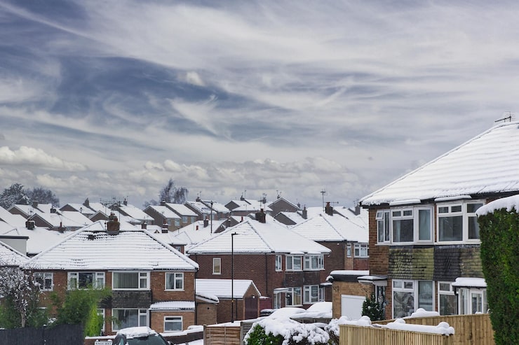Snow-covered suburban houses with white rooftops line a residential neighborhood under a cloudy sky, hinting at the importance of roof lifespan in enduring harsh winters. Green shrubs and fences are visible in the foreground.