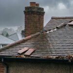 A close-up of a sloped house roof with loose and missing tiles, wet from rain. Leaves are collected in the gutter, and a brick chimney is visible. Other nearby houses and trees appear in the background under a cloudy sky.