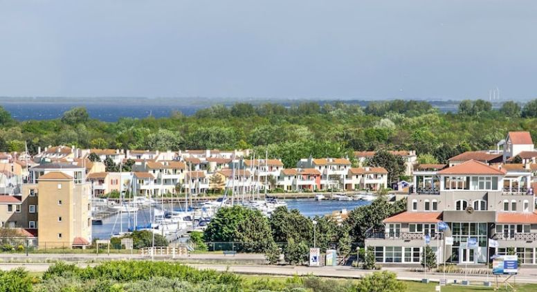 A marina with sailboats docked, surrounded by modern houses and buildings with red and tan roofs, set against a backdrop of green trees and a distant body of water under a cloudy sky.