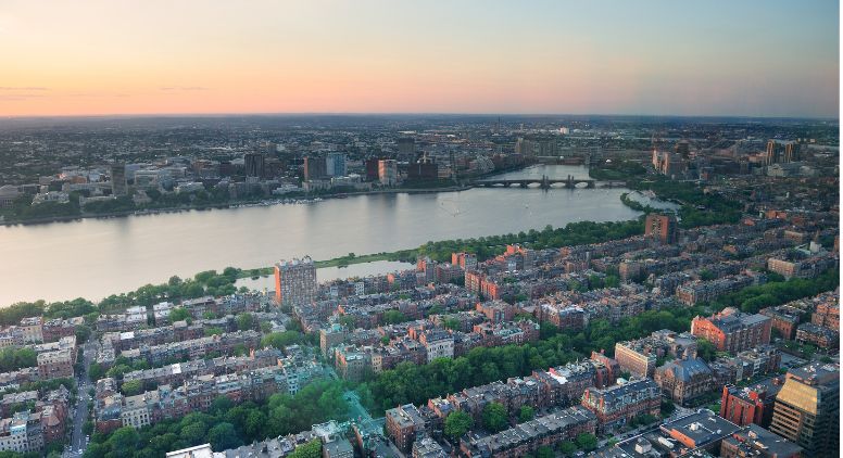 Aerial view of a city at sunset, showing a river, bridge, and densely packed buildings with green trees lining the streets.