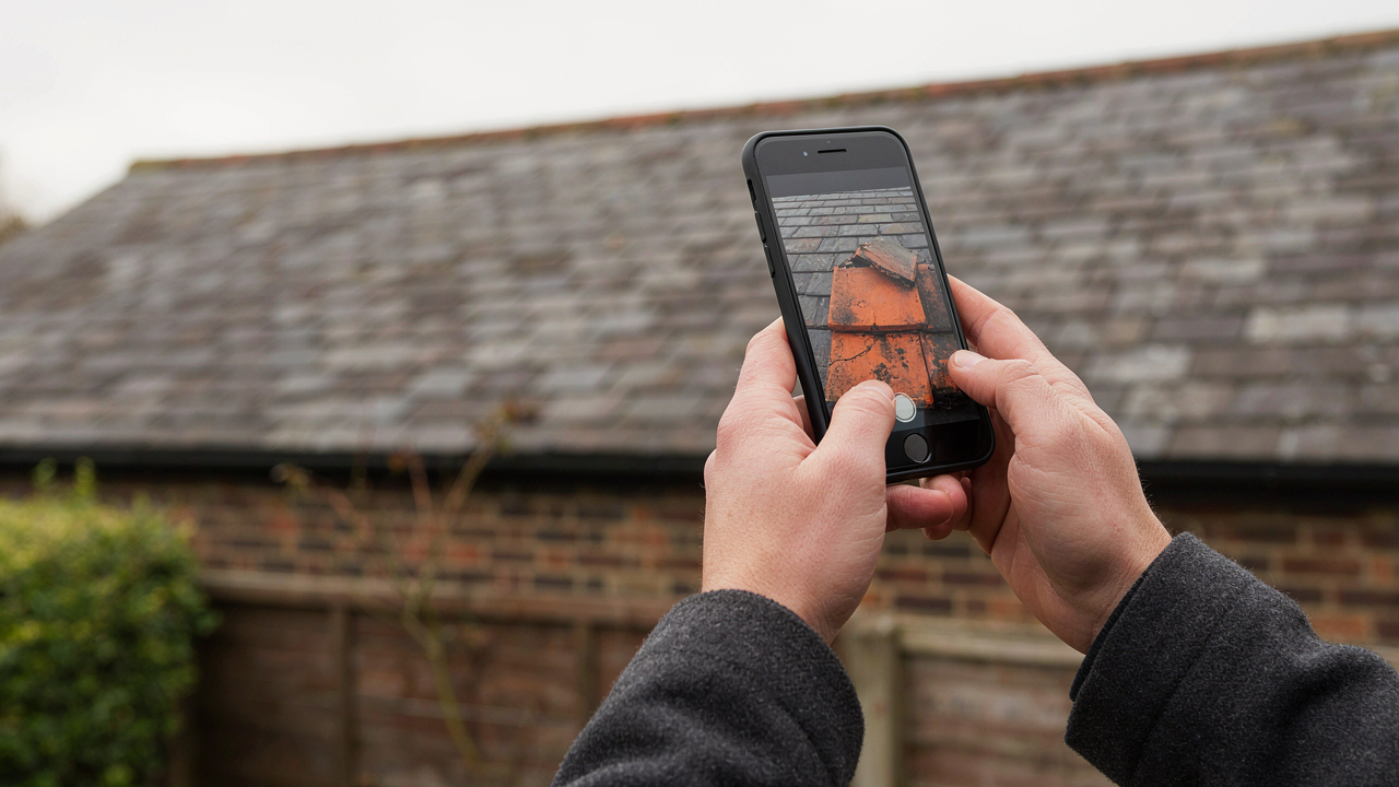 Close-up of hands holding a smartphone, photographing a damaged roof tile on a UK tiled roof.