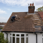 Two workers in safety gear repair and replace roof tiles on a house. Part of the roof is stripped, exposing wooden beams. The neighborhood has similar houses and trees, with a partly cloudy sky above.