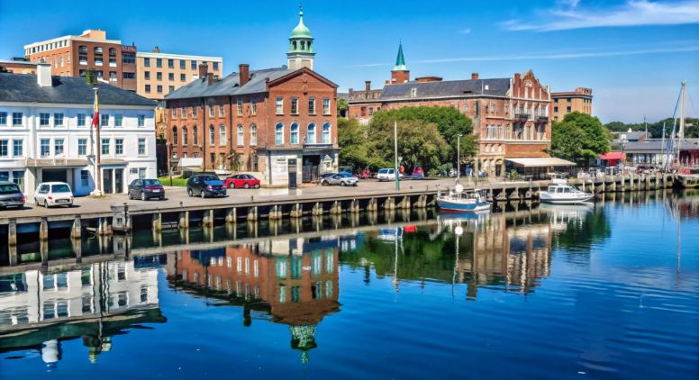 Colorful historic buildings line a waterfront with boats docked and clear blue sky reflected in the calm water, creating a picturesque and peaceful harbor scene.