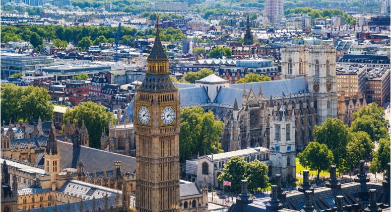 Aerial view of London featuring the Big Ben clock tower and Westminster Abbey, surrounded by green trees and city buildings under a clear sky.